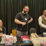 Three people sit behind a table covered in copies of Haverin, Gallus, and Thirty Years of Rain, our three anthologies, plus tablet, tote bags, etc. Sat down is Heather Valentine in strawberry blonde pigtails, while standing is Peter Morrison, a bearded man wearing glasses and a tiara and Brian Milton, also a bearded man with glasses, opening a bottle of Irn Bru.